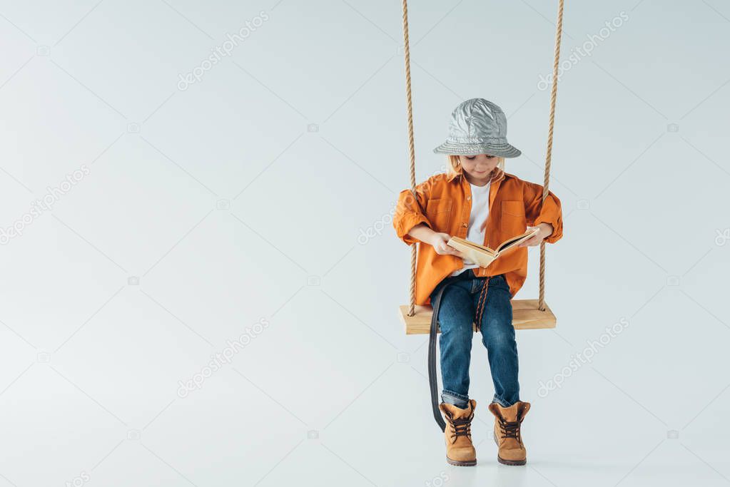 cute kid in jeans and orange shirt sitting on swing and reading book on grey background