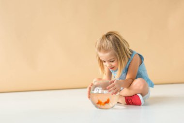 adorable kid sitting with crossed legs and looking at fishbowl on beige background  clipart