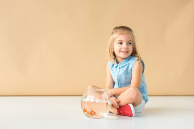 cute kid sitting on floor with fishbowl and looking away on beige background  clipart