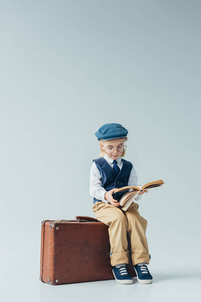 ребенок в ретро жилете и кепке сидит на чемодане и читает книгу на сером фоне
