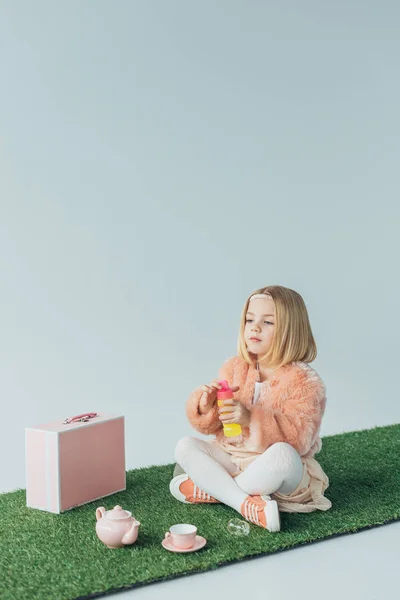 cute kid with crossed legs sitting on grass rug and holding bottle with soap bubbles isolated on grey