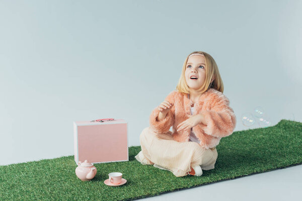 cute kid with crossed legs sitting on grass rug and looking at soap bubbles isolated on grey