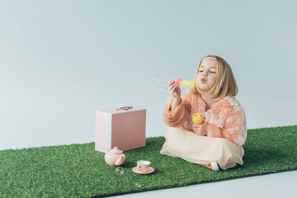 cute child with crossed legs sitting on grass rug and blowing soap bubbles isolated on grey
