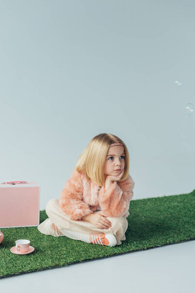 thoughtful child with crossed legs sitting on grass rug and looking away isolated on grey