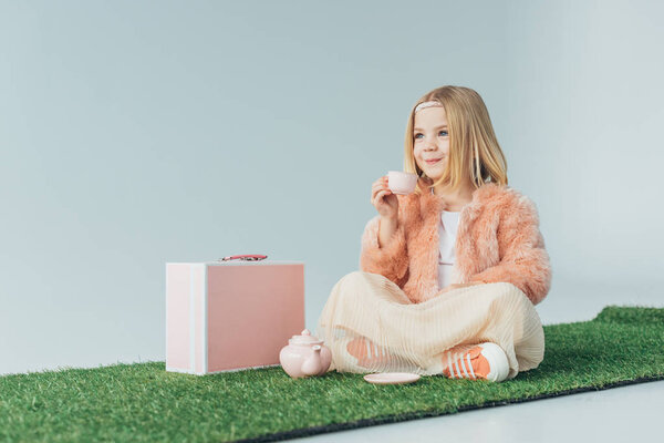 smiling child sitting with crossed legs playing with pink toy dishes isolated on grey