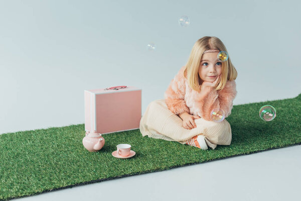 dreamy child with crossed legs sitting on grass rug and looking at soap bubbles isolated on grey