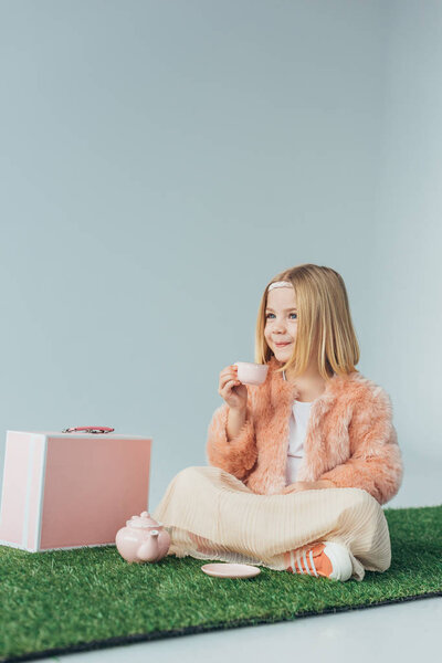 smiling child sitting with crossed legs playing with toy dishes and looking away isolated on grey