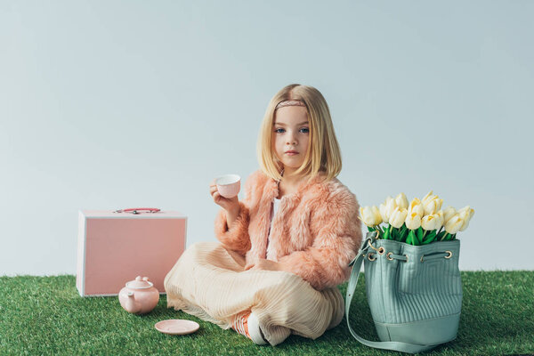 child with crossed legs playing with toy dishes and looking at camera isolated on grey 