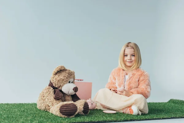 smiling child with crossed legs sitting on grass rug with teddy bear isolated on grey