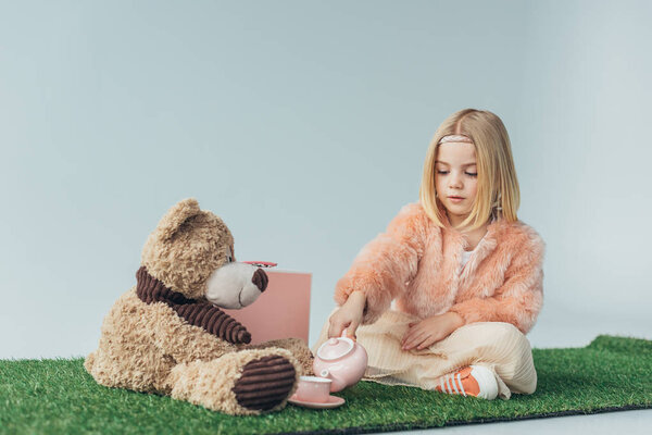 child with crossed legs playing with toy dishes and teddy bear isolated on grey 