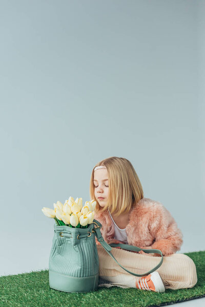 cute kid with crossed legs smelling white tulips in bag isolated on grey