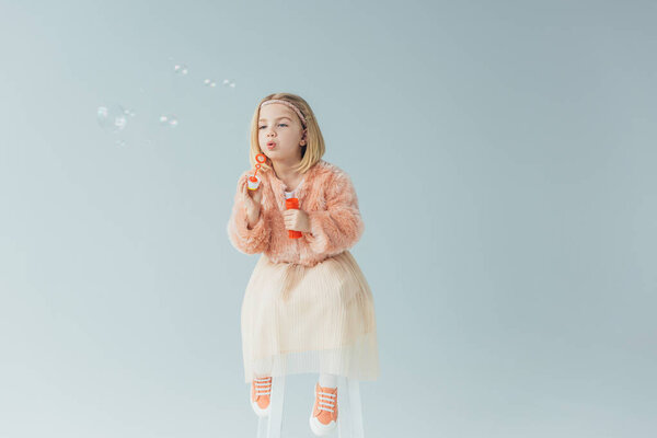 adorable kid in faux fur coat and skirt sitting on highchair and blowing soap bubbles isolated on grey