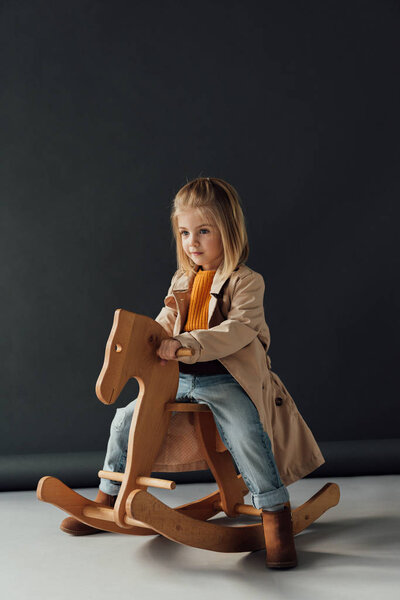  child in trench coat and jeans sitting on rocking horse on black background