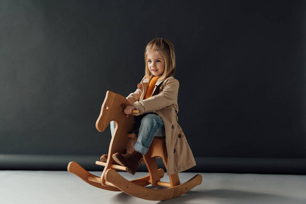  smiling child in trench coat and jeans sitting on rocking horse on black background