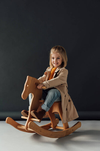happy child in trench coat and jeans sitting on rocking horse on black background