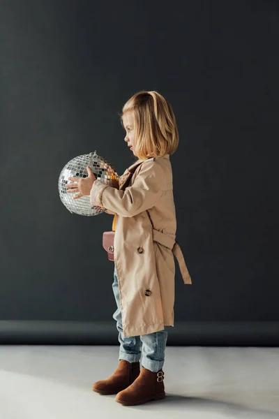 side view of child in trench coat holding disco ball on black background