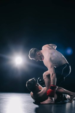 shirtless strong mma fighter in boxing gloves standing above opponent and punching him in head while sportsman lying on floor clipart