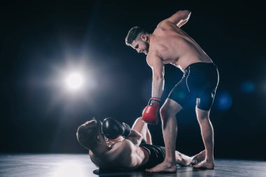 shirtless strong mma fighter in boxing gloves standing above opponent while sportsman lying on floor clipart