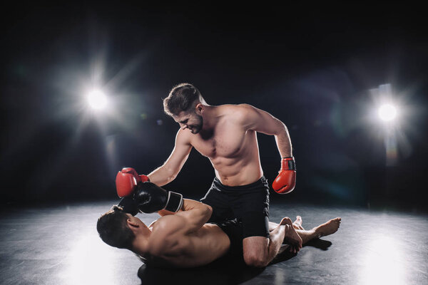 strong mma fighter in boxing gloves punching opponent while sportsman lying on floor