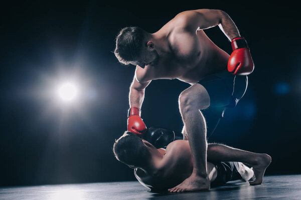 bottom view of strong mma fighter in boxing gloves punching opponent in head while sportsman lying on floor