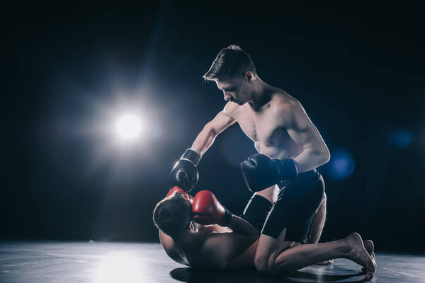 shirtless strong mma fighter in boxing gloves standing on knees above opponent and punching him