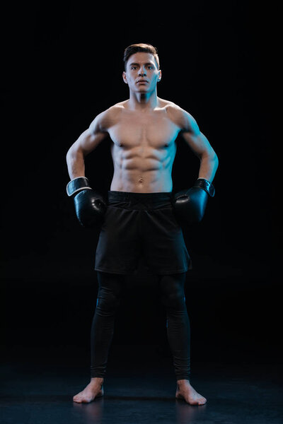 confident muscular barefoot boxer in boxing gloves posing on black
