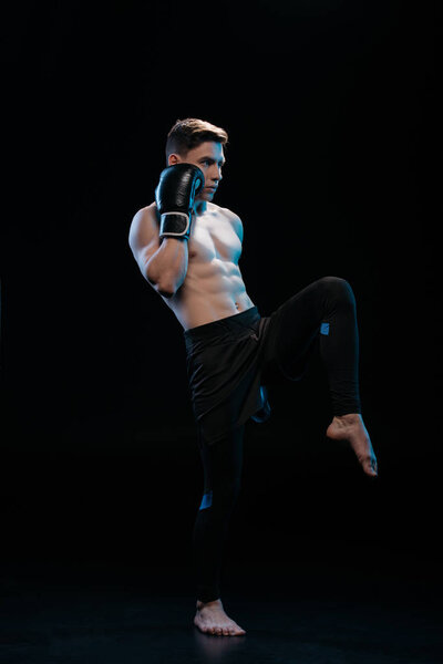 muscular barefoot mma fighter in boxing gloves doing kick on black