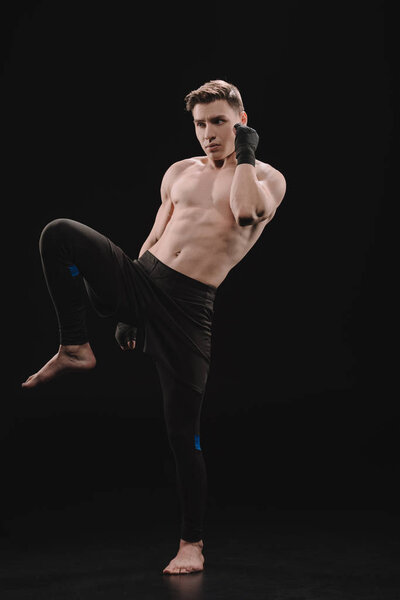 athletic muscular barefoot sportsman in bandages doing kick on black