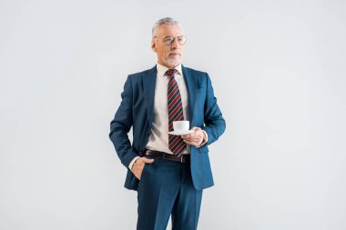 fincan ile fincan tabağı tutarak ve gri izole cebinde el ile ayakta ciddi olgun iş adamı