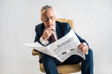 mature businessman sitting in armchair and reading business newspaper isolated on grey clipart