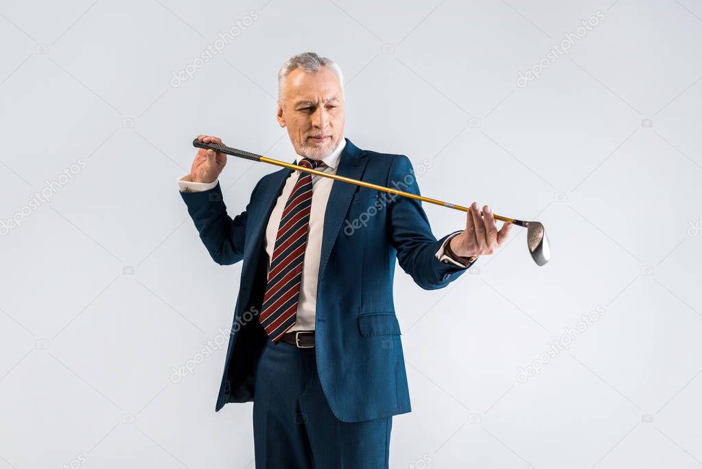 concentrated mature man in suit looking at golf club isolated on grey