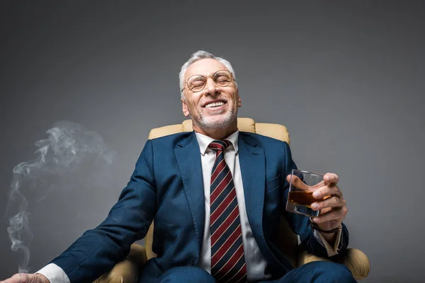 ウィスキーのグラスを押しながらグレーの上に椅子に座っているスーツで笑顔の成熟したビジネスマン — ストック写真
