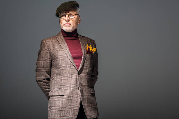 trendy mature man in cap and glasses standing on grey