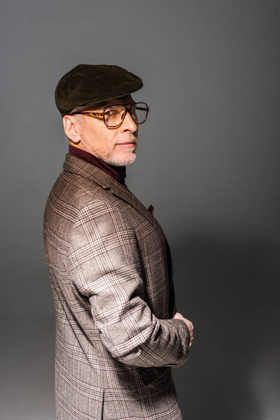 serious and stylish mature man in cap and glasses on grey