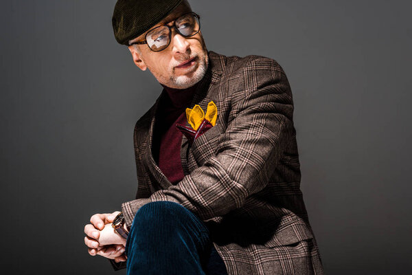 trendy and pensive mature man in cap and glasses on grey 