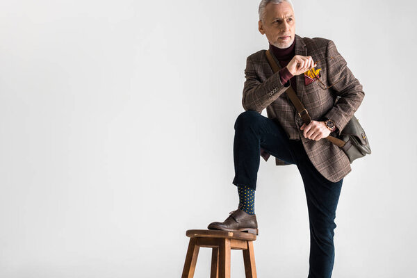 stylish mature man putting leg on chair while standing and holding glasses on grey 