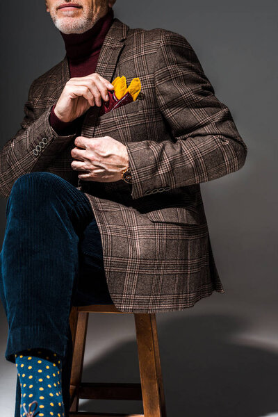 cropped view of fashionable mature man sitting on chair and touching handkerchief on grey 
