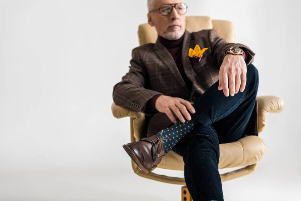 selective focus of mature man in glasses sitting in armchair with crossed legs on grey 