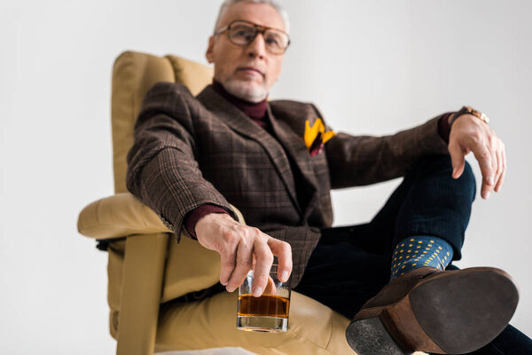 selective focus of mature man sitting in armchair with crossed legs and holding glass of whiskey isolated on grey
