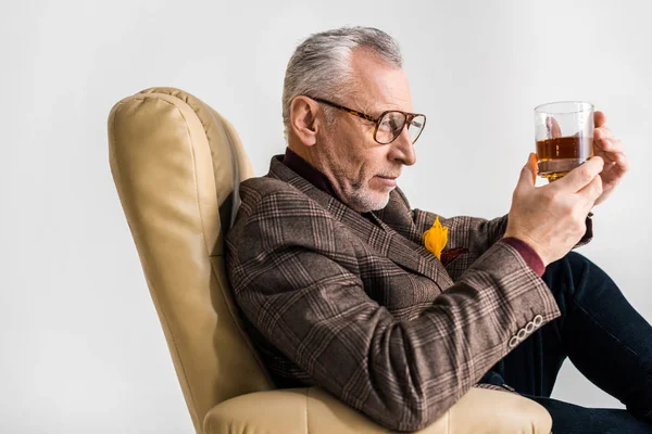 Зрелый Мужчина Сидит Кресле Смотрит Стакан Виски Изолированный Сером — стоковое фото
