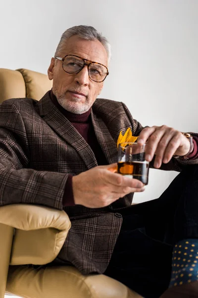 stylish mature man sitting in armchair and holding glass of whiskey isolated on grey