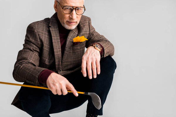 stylish middle aged man in glasses holding golf club and while sitting isolated on grey