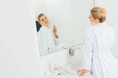 diş fırçası tutan ve aynaya bakarak beyaz bornoz içinde kadın seçici odak 