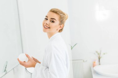 smiling woman in white bathrobe holding cosmetic cream and looking at camera in bathroom clipart