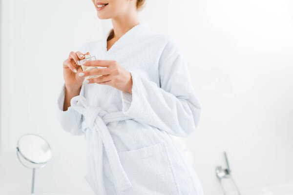 обрезанный вид женщины в белом халате держа духи в ванной комнате
 