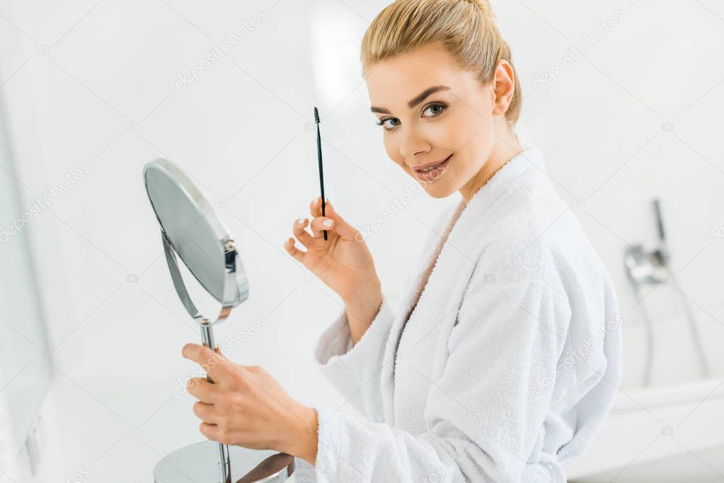 attractive woman in white bathrobe holding cosmetic brush and looking at camera