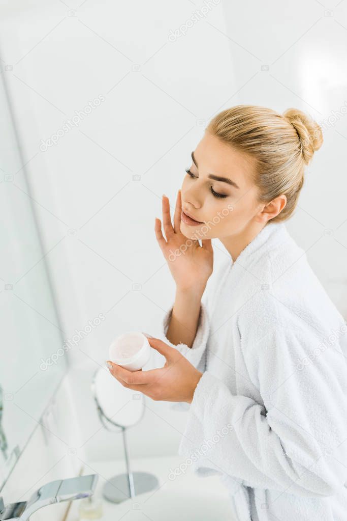 attractive and blonde woman in white bathrobe applying face cream in bathroom