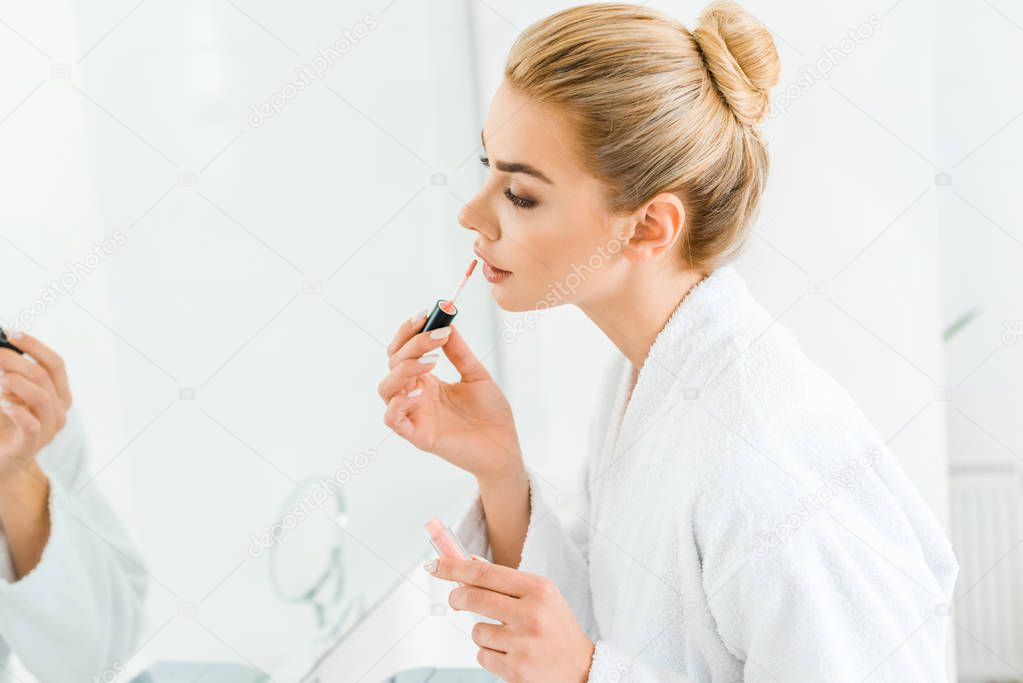selective focus of beautiful woman in white bathrobe applying lip gloss in bathroom 