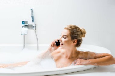 köpüklü banyo ve banyoda smartphone cep telefonu ile konuşurken çekici ve gülümseyen kadın 