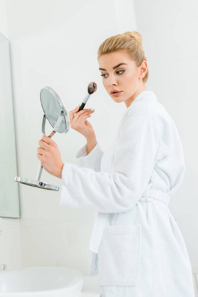 attractive and blonde woman in white bathrobe using cosmetic brush in bathroom 